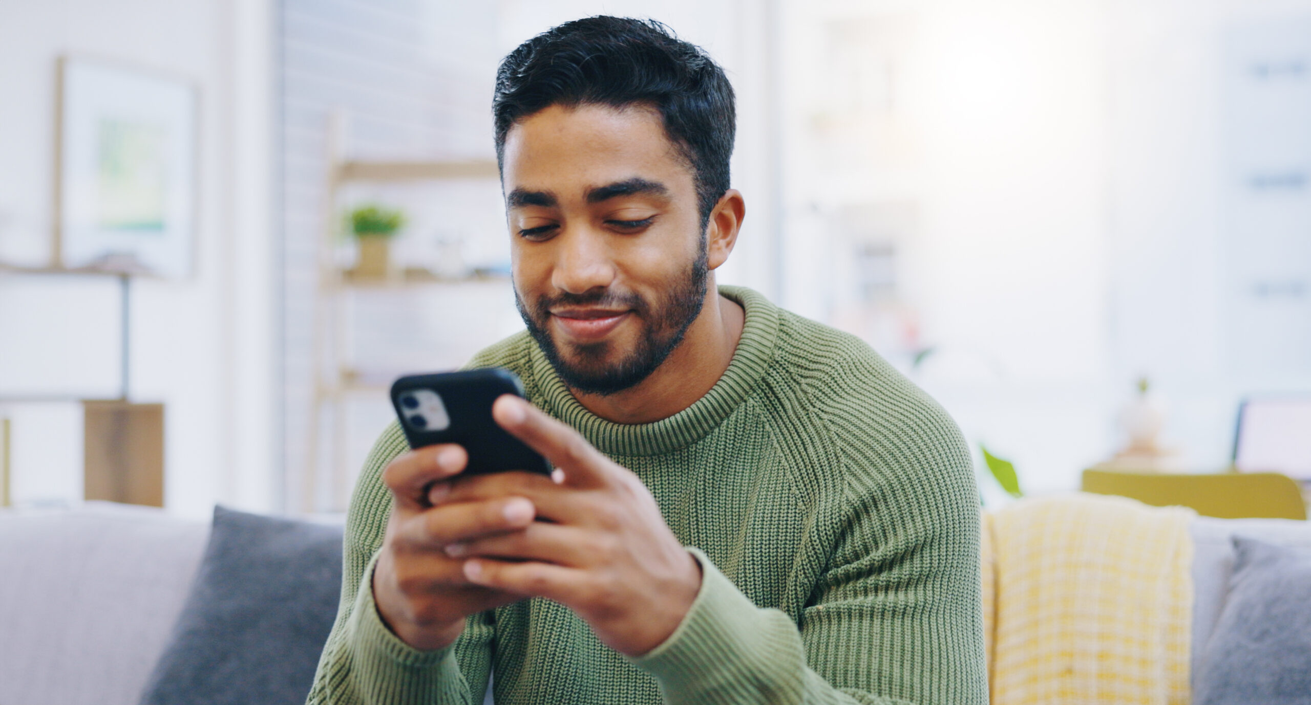 Man, living room couch and texting with phone, smile and click for contact, meme and notification in home. Person, smartphone and happy for scroll, reading and app for social network, video and relax.